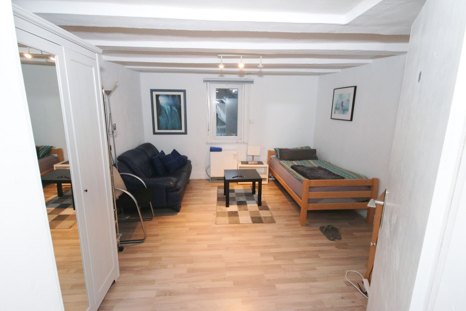 Einzelzimmer mit Bett, Tisch, Couch einem Schrank sowie mit NAchttisch auf dem eine Lampe steht und Bildern an der Wand sowie einem Fenster.